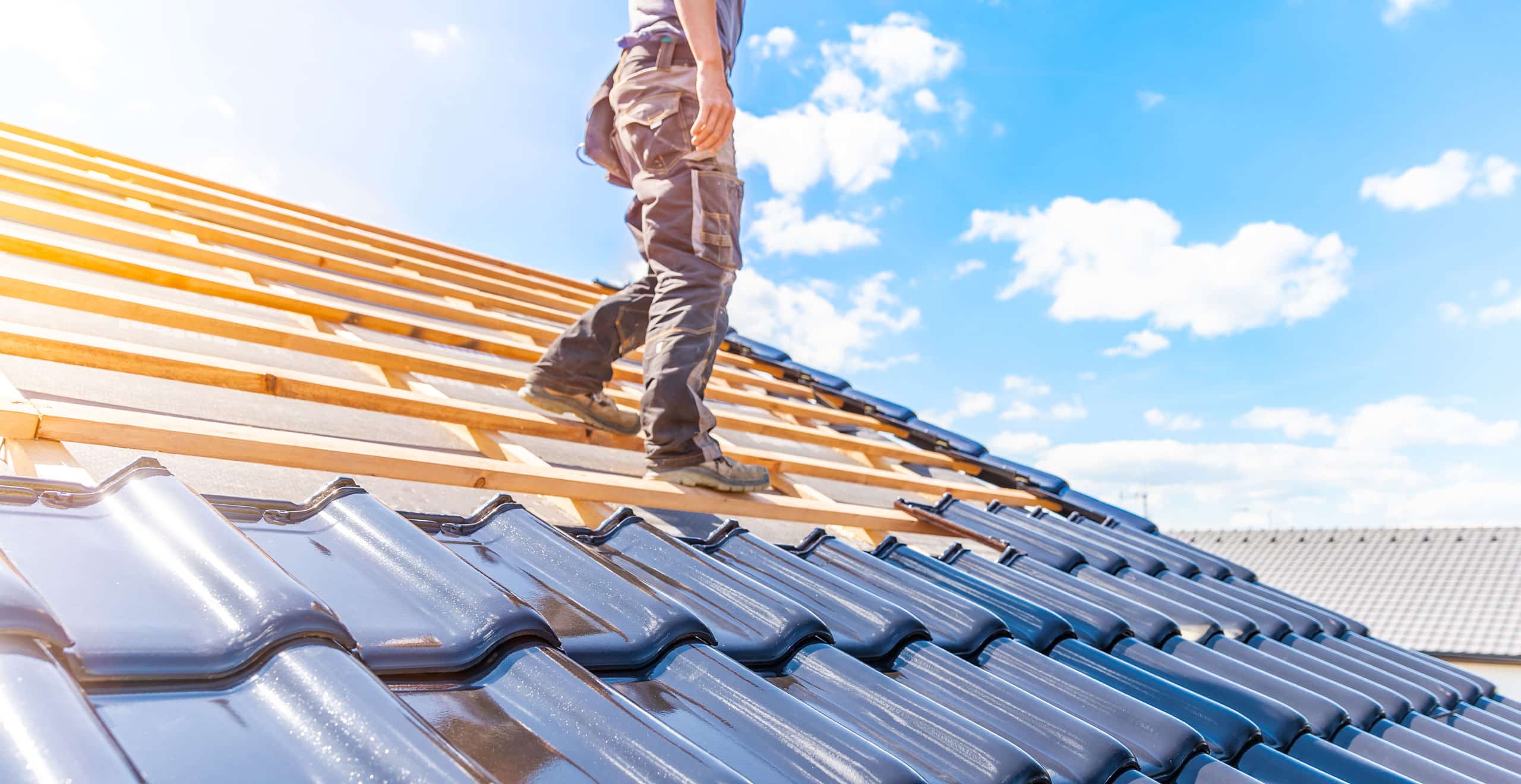 Dachdecker steht auf Dach mit schwarzen Ziegeln. Sonne und blauer Himmel im Hintergrund.