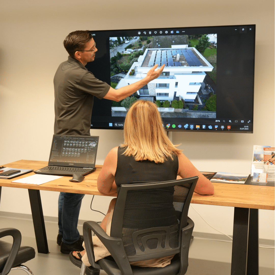 Mann zeigt Frau auf großem Bildschirm ein Haus mit Photovoltaik.
